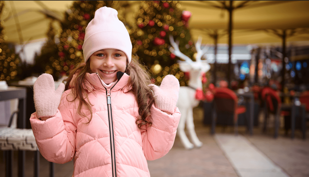 Smiling young girl in a pink winter coat, hat, and mittens stands outdoors near blurred Christmas decorations including lights and a white reindeer figure.