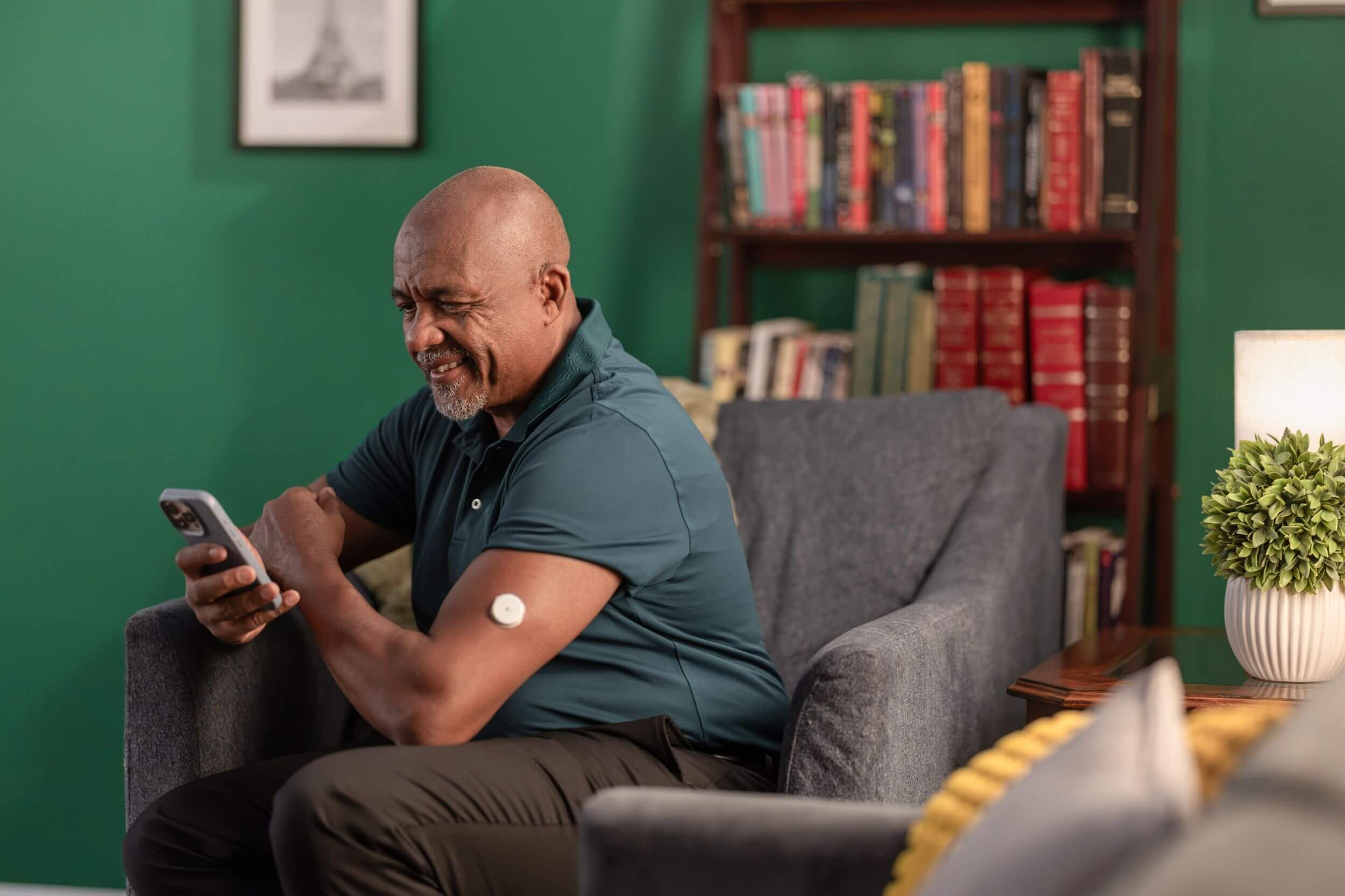 cfhp-medicare-2 An elderly man sits on a comfy grey sofa, smiling at his smartphone, with a round sensor patch on his upper arm and wooden bookshelf behind him.