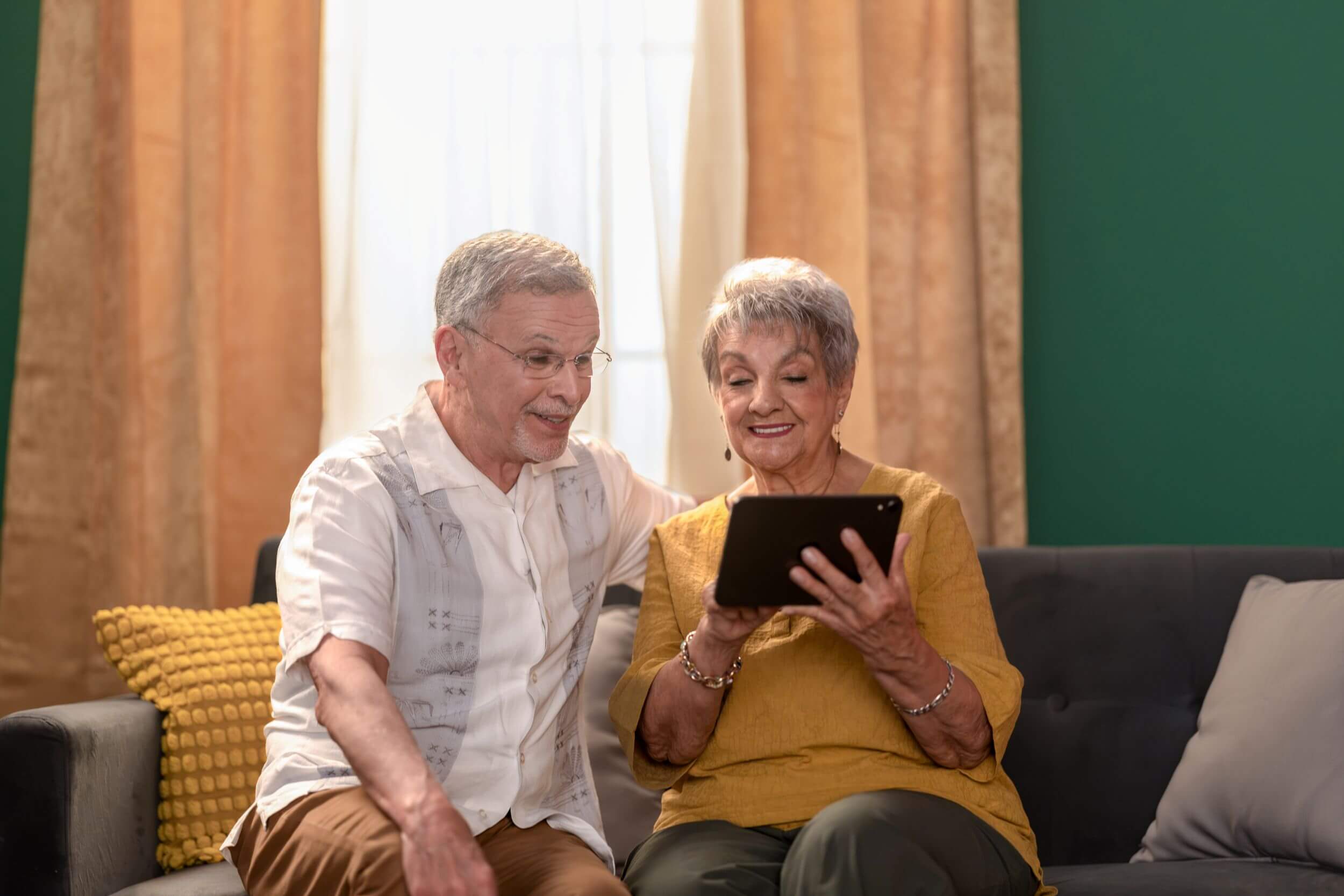 Senior couple sitting on a grey sofa, smiling while looking together at a black tablet.